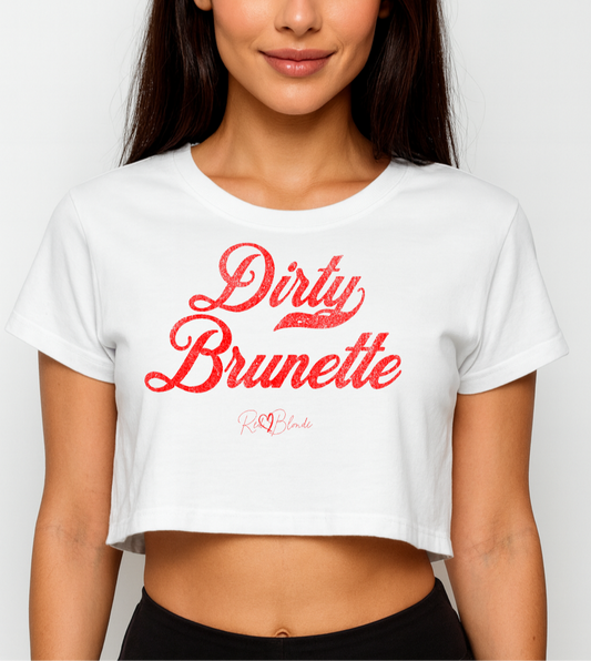 brunette wears a white women’s crop top featuring the phrase “Dirty Brunette” in bold, red milkstore vintage-style script across the chest. Below the phrase, the brand name “RedBlonde” is printed in a smaller, elegant handwritten font.