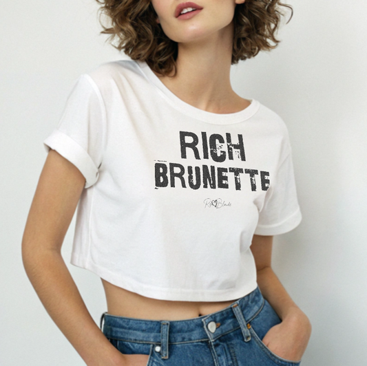 brunette wears a white crop top with a relaxed fit, short sleeves, and a wide crew neckline. The front features bold black distressed Ed Gein lettering reading “RICH BRUNETTE” with the RedBlonde signature logo underneath in black
