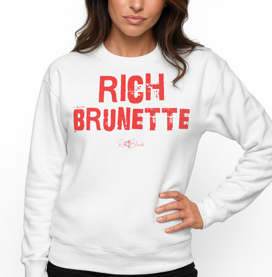 Brunette wears a white unisex premium sweatshirt with bold distressed red Ed Geinitext “RICH BRUNETTE” on the front, featuring a subtle RedBlonde logo beneath the lettering and on the sleeve cuff and inside neck