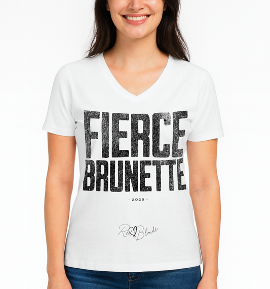 brunette wears a white women’s V-neck T-shirt featuring the bold black distressed text “FIERCE BRUNETTE” in large North Road capital letters across the chest. Beneath the text, the year “2025” is printed in small type, with the “RedBlonde” brand signature logo below it. The inner collar shows the “RedBlonde” brand name in red along with sizing and care details, and “RedBlonde” is also printed in small red letters on the right sleeve.