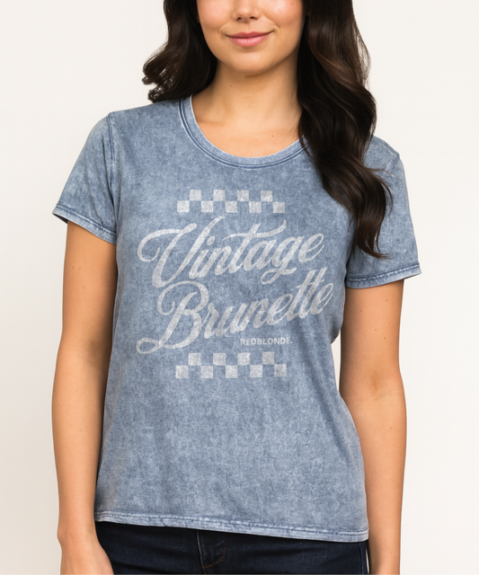 brunette wears a blue acid wash short-sleeve T-shirt featuring the bold red Milkstore 1 script text “Vintage Brunette” across the chest with checkerboard accents above and below the lettering. The brand name “RedBlonde.” is printed in small capital letters beneath the word “Brunette.” The shirt has a casual, slightly distressed look with visible stitching along the seams. Branded RedBlonde on inside collar and right sleeve