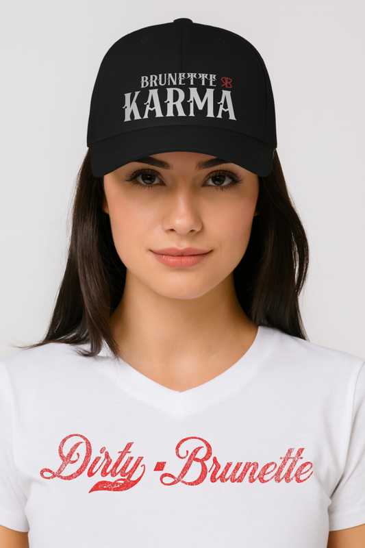 brunette wears a black retro trucker cap with mesh back and a Dirty Brunette white v-neck t-shirt. The front features bold white embroidered text reading "BRUNETTE KARMA" with a small red RB logo to the right.