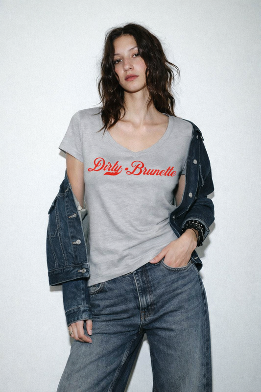 brunette wears a grey V-neck T-shirt laid flat on a white background. It has short sleeves and a red Milkstore 01 script graphic across the chest reading “Dirty-Brunette.” A small RedBlonde label is visible at the inside back neck.