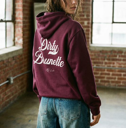 Brunette model wears a plum coloured hoodie with bold white Milkstore vintage-style script text “Dirty Brunette” across the back and a subtle RedBlonde signature logo underneath.