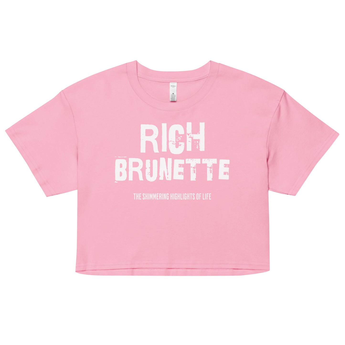A bubblegum women’s crop top with a relaxed fit, short sleeves, and a wide crew neckline. The front features bold white distressed lettering reading “RICH BRUNETTE” with the tagline “THE SHIMMERING HIGHLIGHTS OF LIFE” in smaller text below.