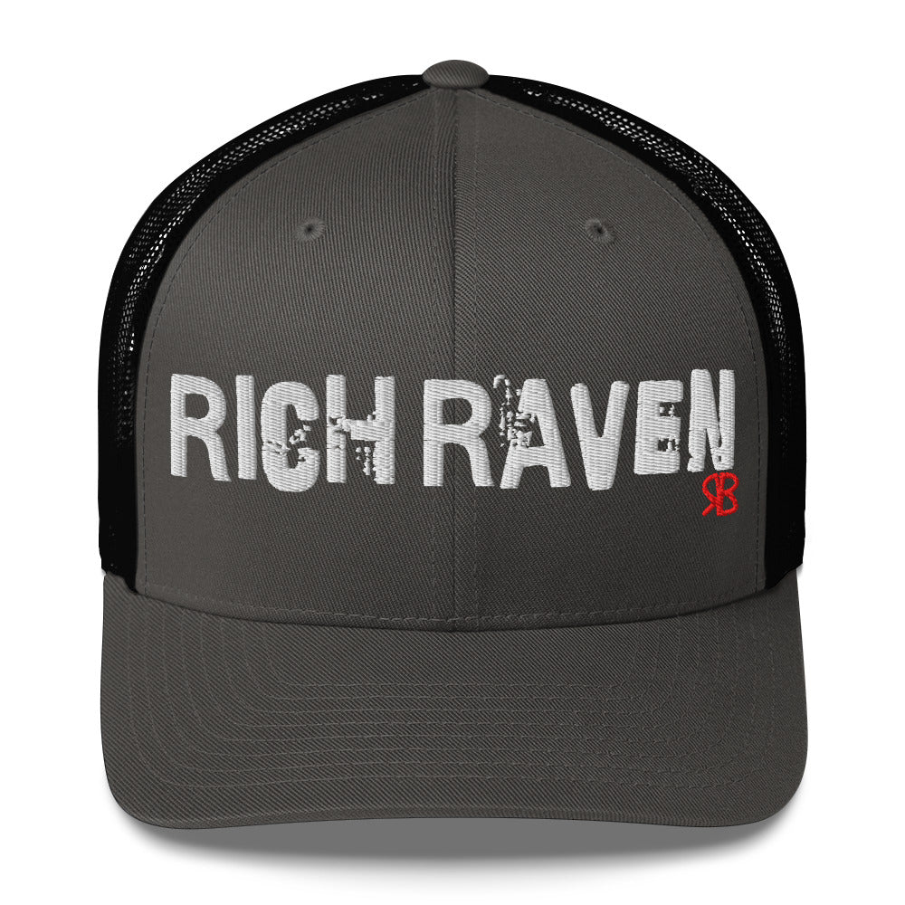 charcoal/black mesh-back trucker cap with the bold distressed text “RICH RAVEN” embroidered in white across the front, and a small red “RB” logo detail beneath the text. The cap has a structured front panel and curved brim
