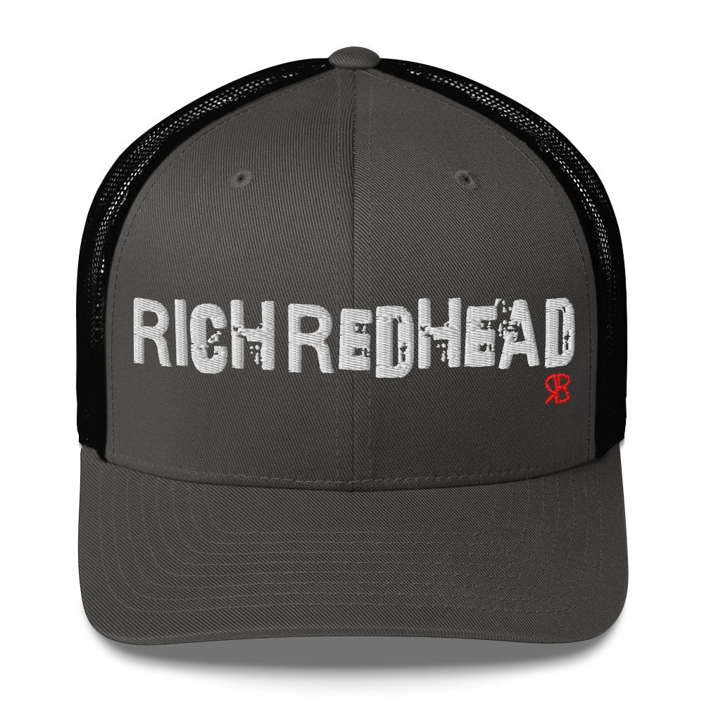 charcoal/black trucker cap with the bold distressed text “RICH REDHEAD” embroidered in white across the front, and a small red “RB” logo detail beneath the text. The cap has a structured front panel and curved brim