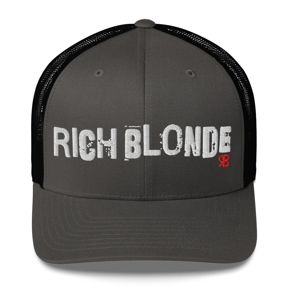 charcoal grey/Black mesh-back trucker cap with the bold distressed text “RICH BLONDE” embroidered in white across the front, and a small red “RB” logo detail beneath the text. The cap has a structured front panel and curved brim