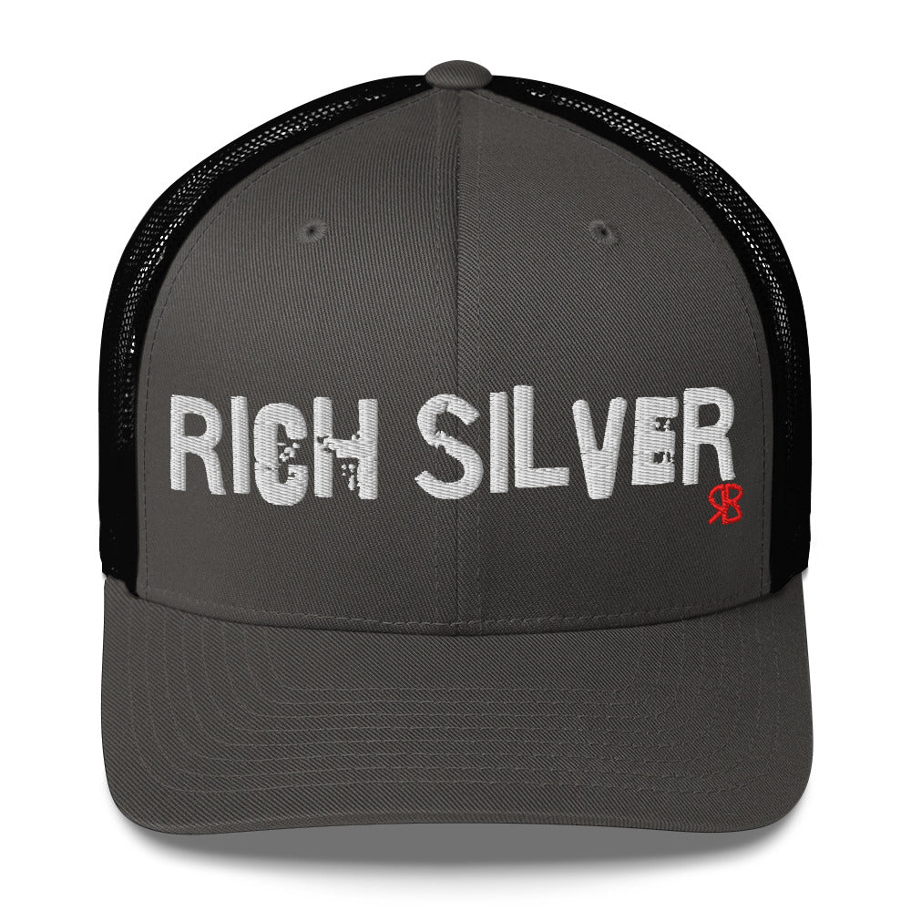 Black/charcoal grey mesh-back trucker cap with the bold distressed text “RICH SILVER” embroidered in white across the front, and a small red “RB” logo detail beneath the text. The cap has a structured front panel and curved brim