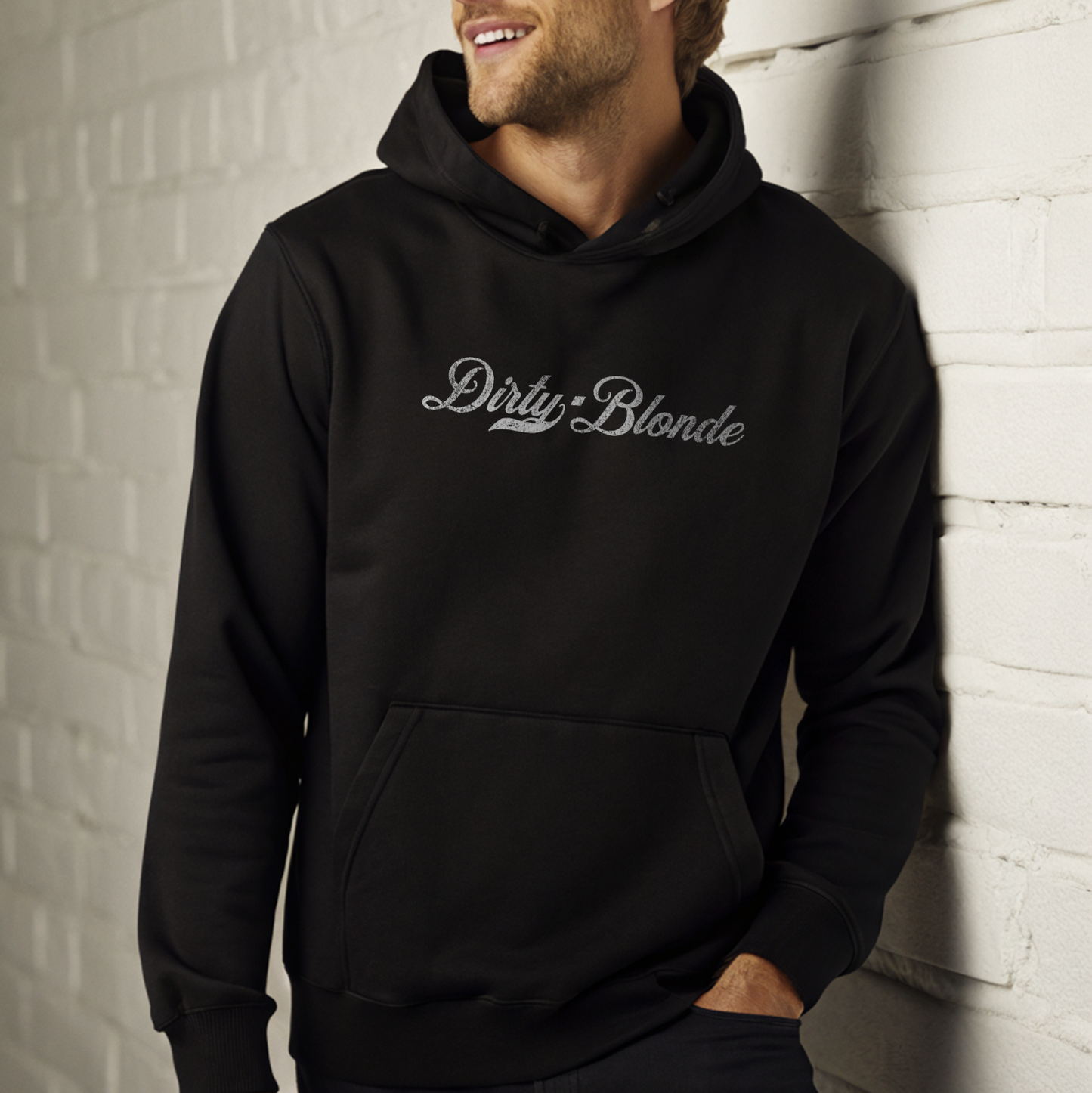 Blonde smiling man leaning on a white wall wearing a classic black relaxed fit unisex hoodie with the words 'dirty Blonde' written in white vintage milkstore font, like coco cola script