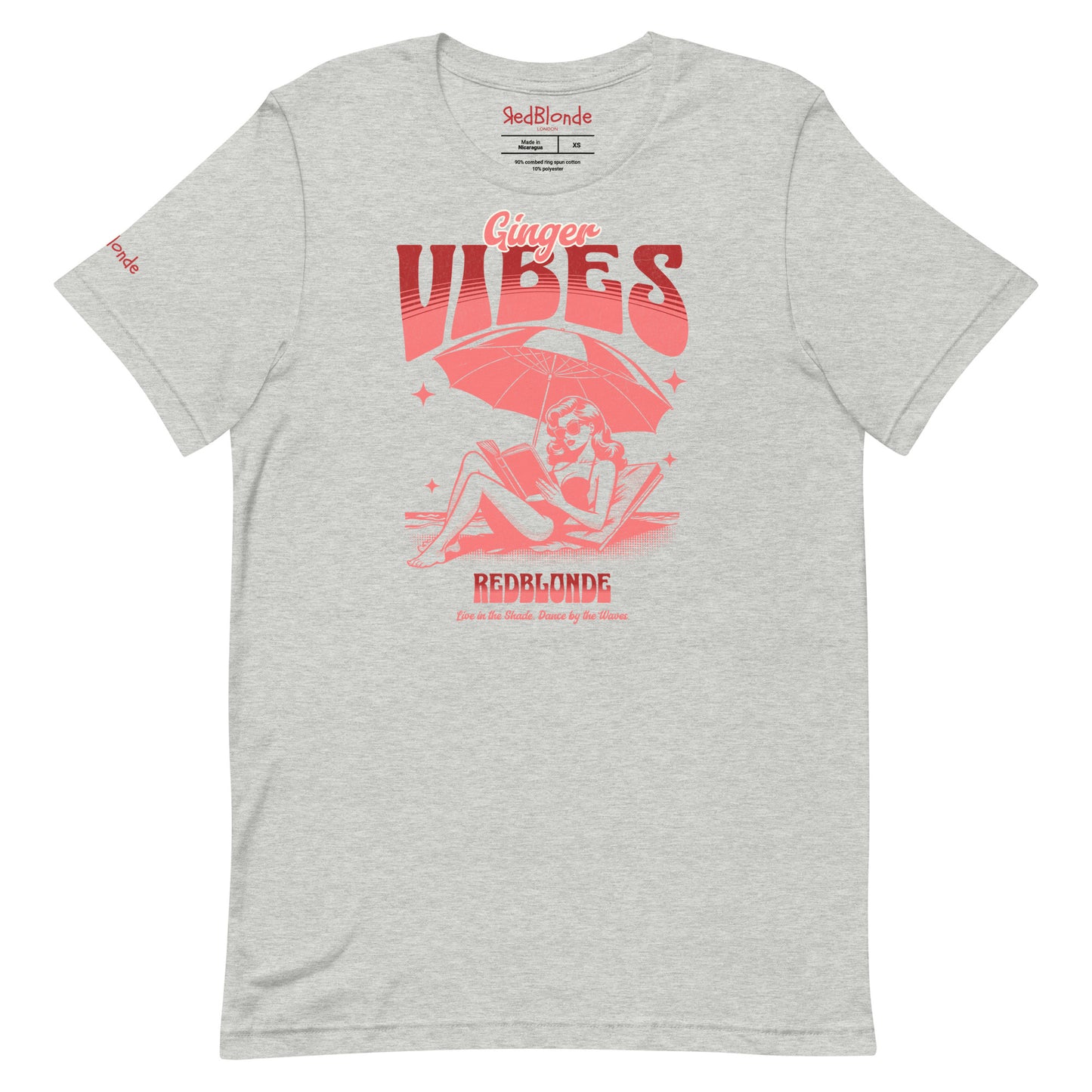 grey unisex t-shirt featuring a retro-style red illustration of a redhead woman lounging under a beach umbrella reading a book, with bold text reading "Ginger Vibes" above and "RedBlonde" below, alongside the tagline "Live in the shade. Dance by the waves." Branded RedBlonde on inside neck and right sleeve