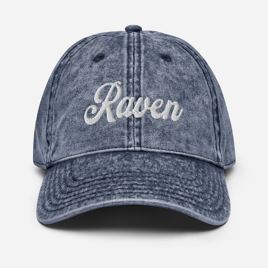flatlay of vintage blue denim wash effect baseball cap with the word 'Raven' embroidered on the front in white adventure type script font