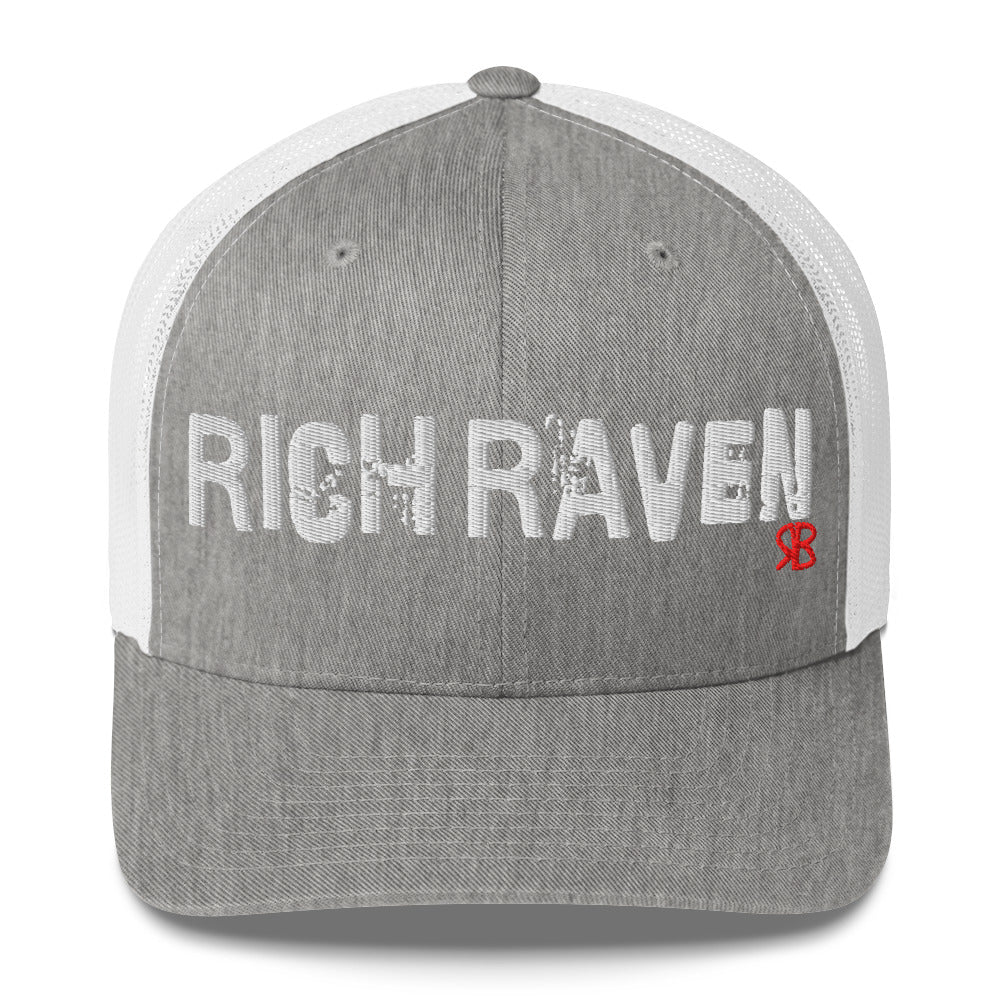 light grey/white mesh-back trucker cap with the bold distressed text “RICH RAVEN” embroidered in white across the front, and a small red “RB” logo detail beneath the text. The cap has a structured front panel and curved brim