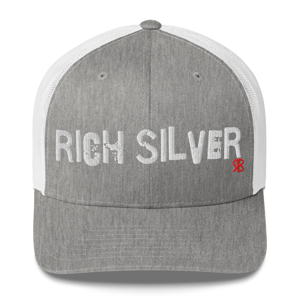 light grey/white mesh-back trucker cap with the bold distressed text “RICH SILVER” embroidered in white across the front, and a small red “RB” logo detail beneath the text. The cap has a structured front panel and curved brim