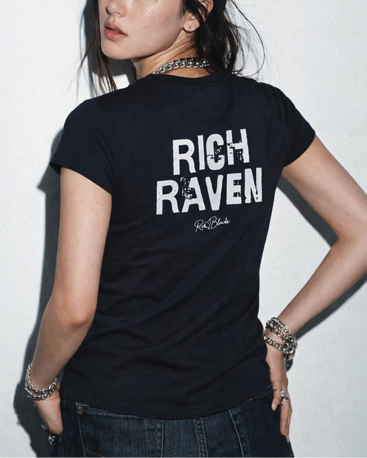 model wears a black slightly fitted women’s henley T-shirt with short sleeves and a gently curved hem. Centered on the upper back is bold distressed black Ed Gein text reading “RICH RAVEN” with a small RedBlonde signature script logo beneath it. The shirt is shown on a plain white background.