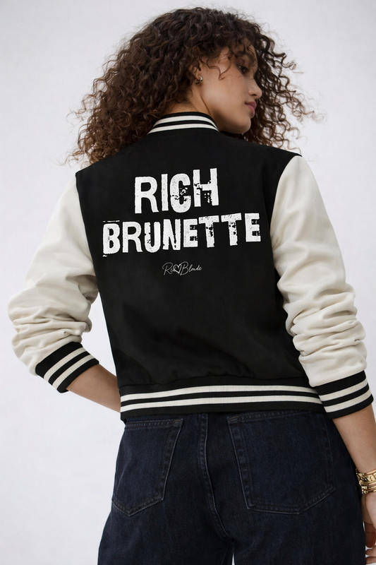 model wears a black & white varsity-style jacket with cream sleeves and striped ribbed trims. The back features large white text reading “RICH BRUNETTE” in Ed Gein font with a small RedBlonde signature logo in the centre between the words