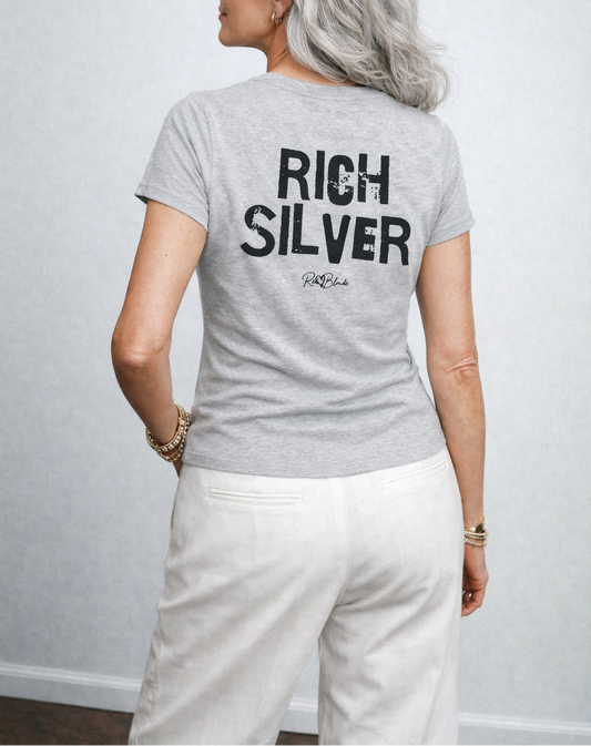 model wears a light grey, slightly fitted women’s henley T-shirt with short sleeves and a gently curved hem. Centered on the upper back is bold distressed black Ed Gein text reading “RICH SILVER,” with a small RedBlonde signature script logo beneath it. The shirt is shown on a plain white background.