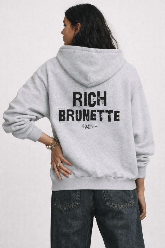 model wears a grey hem to hood zip-up hoodie with cropped oversized fit and ribbed cuffs/hem, featuring large distressed black “RICH BRUNETTE” Ed Gein text across the back with a small “RedBlonde” script logo beneath.