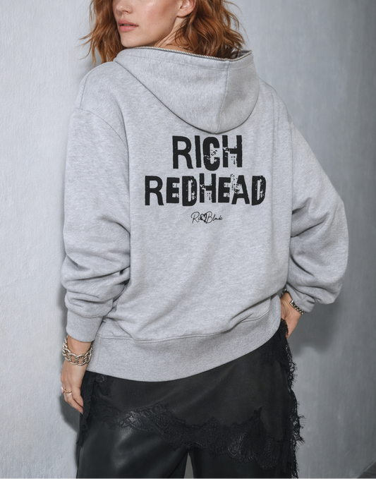 model wears a light grey hem to hood zip-up hoodie with cropped oversized fit and ribbed cuffs/hem, featuring large distressed black “RICH REDHEAD” Ed Gein text across the back with a small “RedBlonde” script logo beneath.