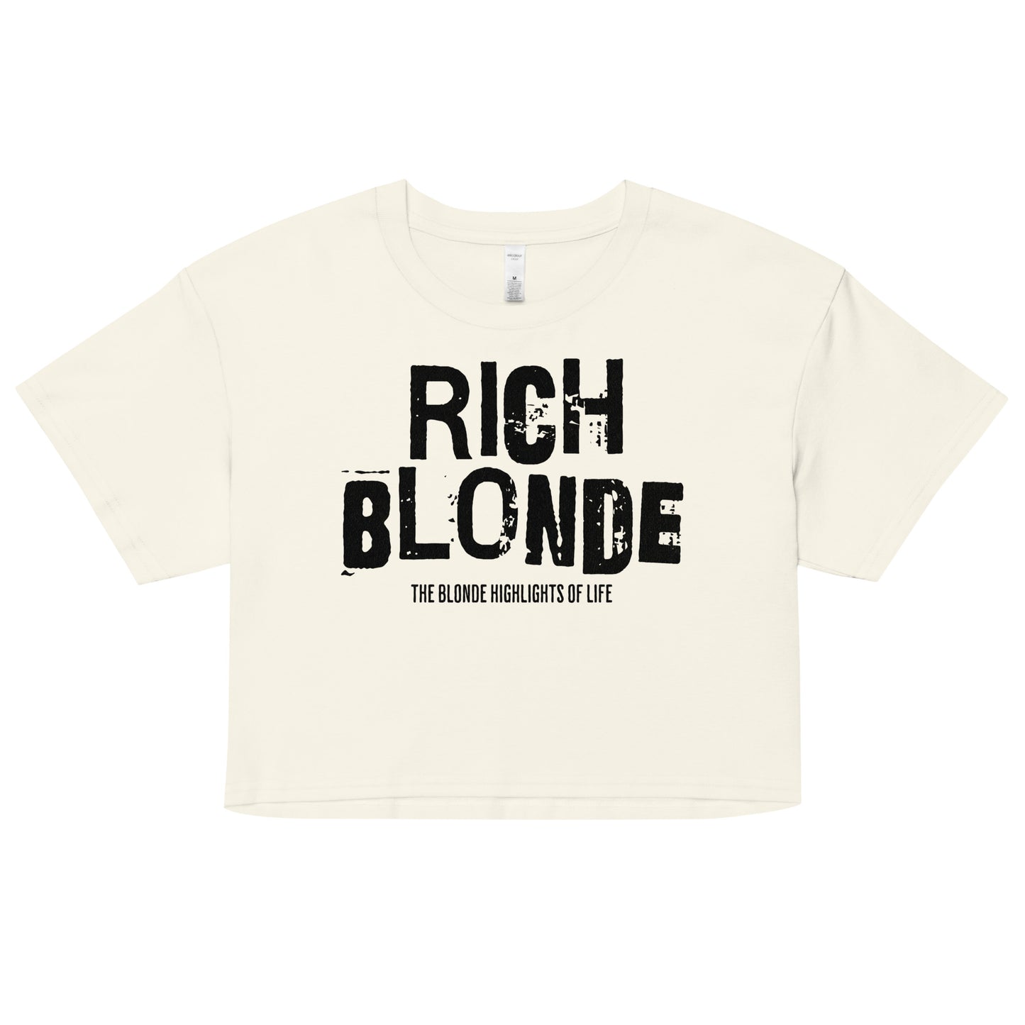 A natural women’s crop top with a relaxed fit, short sleeves, and a wide crew neckline. The front features bold black distressed lettering reading “RICH BLONDE” with the tagline “THE BLONDE HIGHLIGHTS OF LIFE” in smaller text below. The minimalist design contrasts sharply against the soft cream fabric