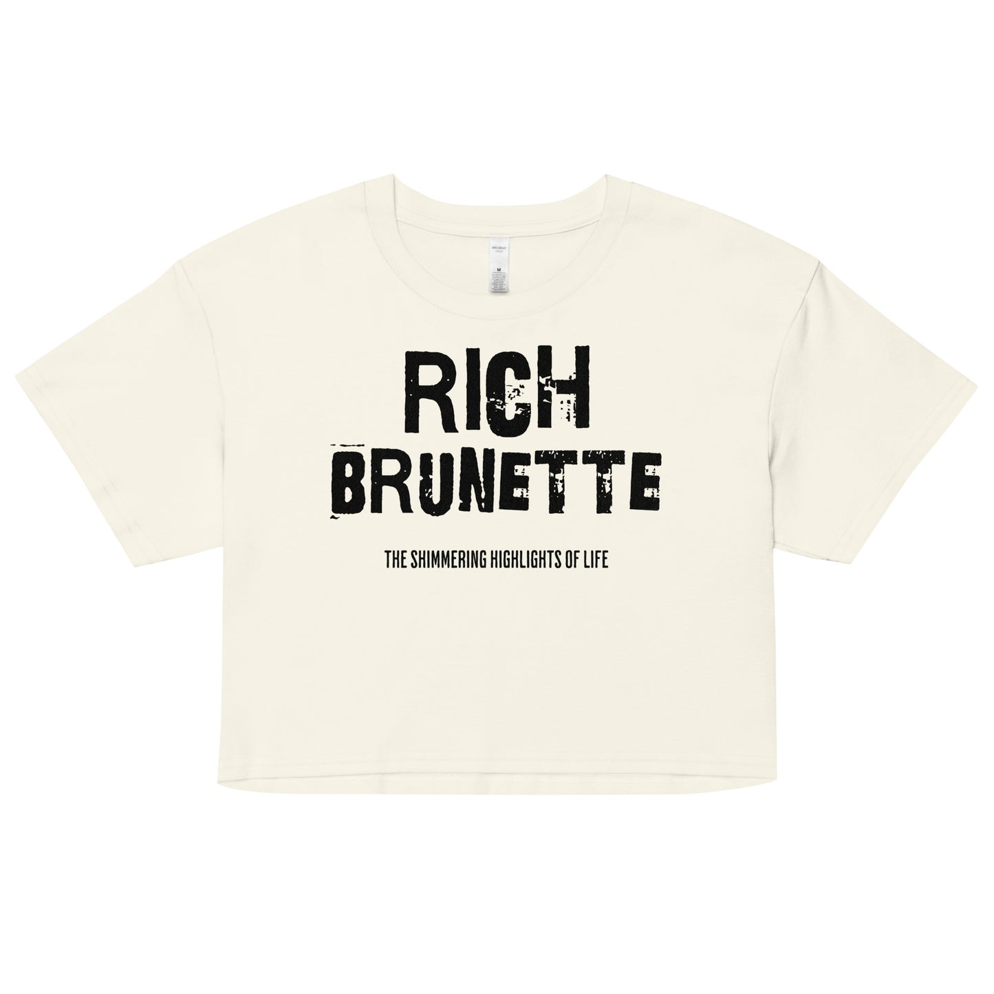 A natural women’s crop top with a relaxed fit, short sleeves, and a wide crew neckline. The front features bold black distressed lettering reading “RICH BRUNETTE” with the tagline “THE SHIMMERING HIGHLIGHTS OF LIFE” in smaller text below. The minimalist design contrasts sharply against the soft cream fabric