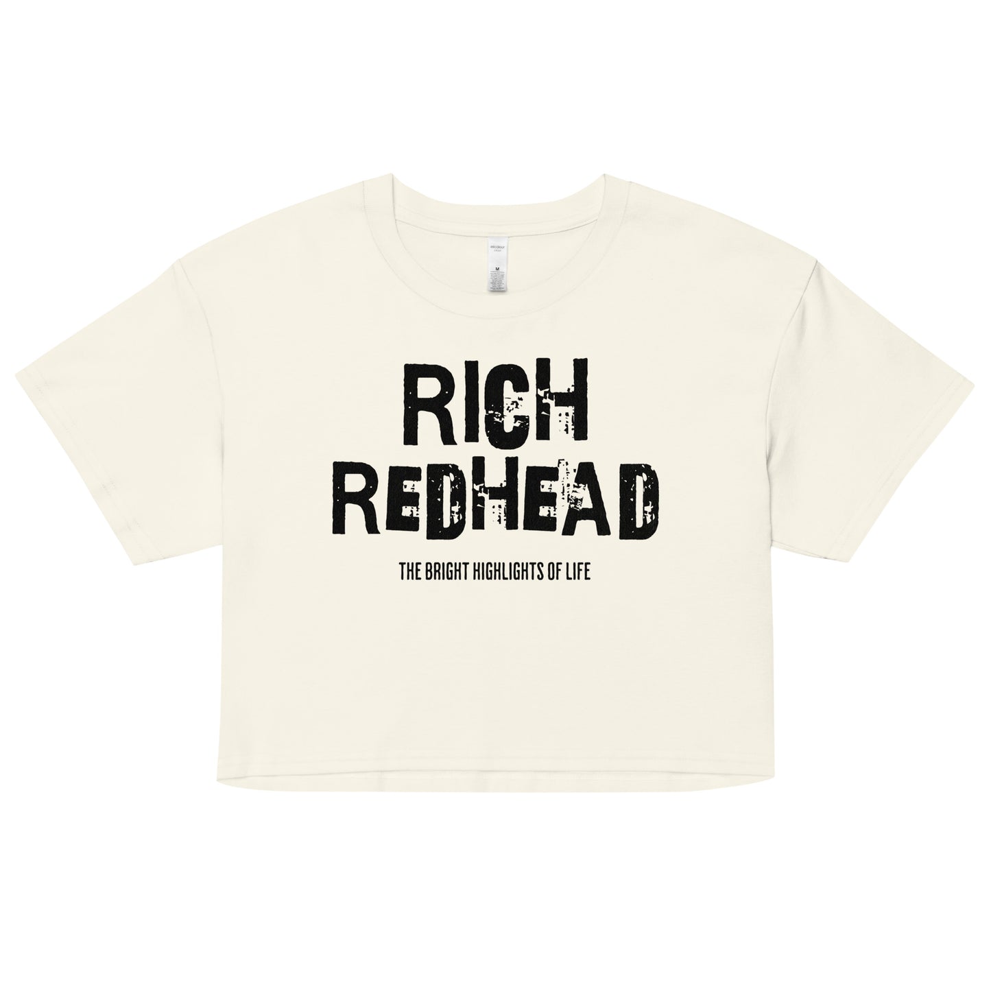 A natural women’s crop top with a relaxed fit, short sleeves, and a wide crew neckline. The front features bold black distressed lettering reading “RICH REDHEAD” with the tagline “THE BRIGHT HIGHLIGHTS OF LIFE” in smaller text below. The minimalist design contrasts sharply against the soft cream fabric