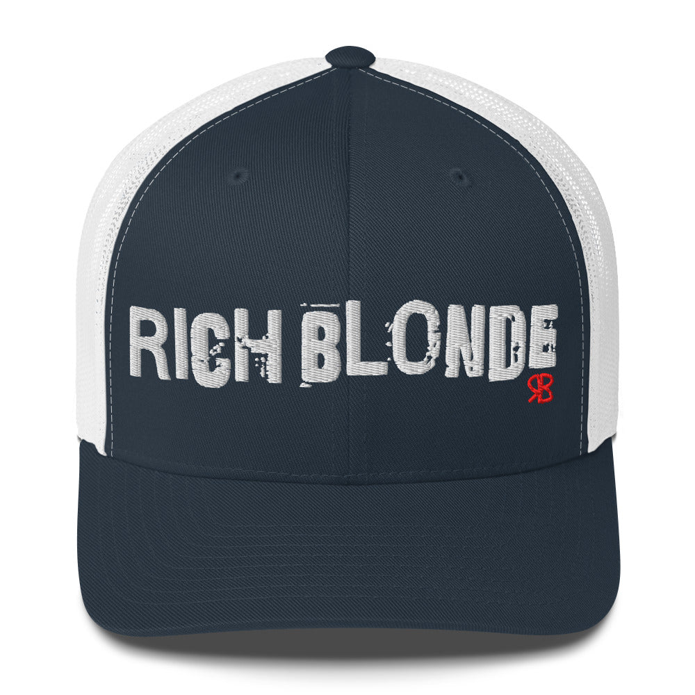 navy/white mesh-back trucker cap with the bold distressed text “RICH BLONDE” embroidered in white across the front, and a small red “RB” logo detail beneath the text. The cap has a structured front panel and curved brim