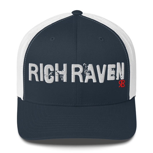 navy/white mesh-back trucker cap with the bold distressed text “RICH RAVEN” embroidered in white across the front, and a small red “RB” logo detail beneath the text. The cap has a structured front panel and curved brim