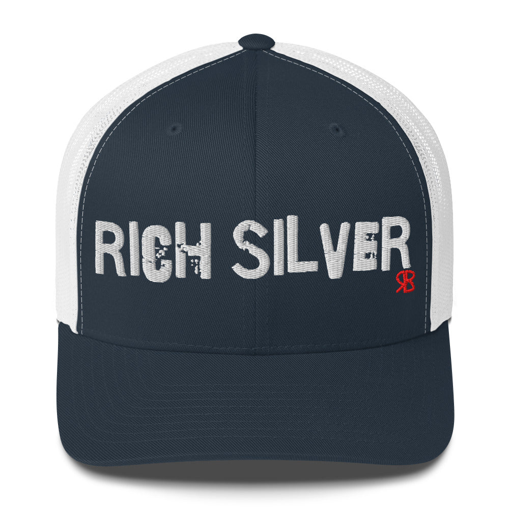 navy/white mesh-back trucker cap with the bold distressed text “RICH SILVER” embroidered in white across the front, and a small red “RB” logo detail beneath the text. The cap has a structured front panel and curved brim