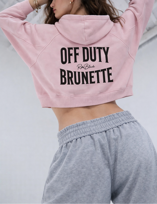 model wears a soft pink cropped hoodie with a hood and long sleeves, featuring large black ‘OFF DUTY BRUNETTE' text and a small RedBlonde logo on the back.”
