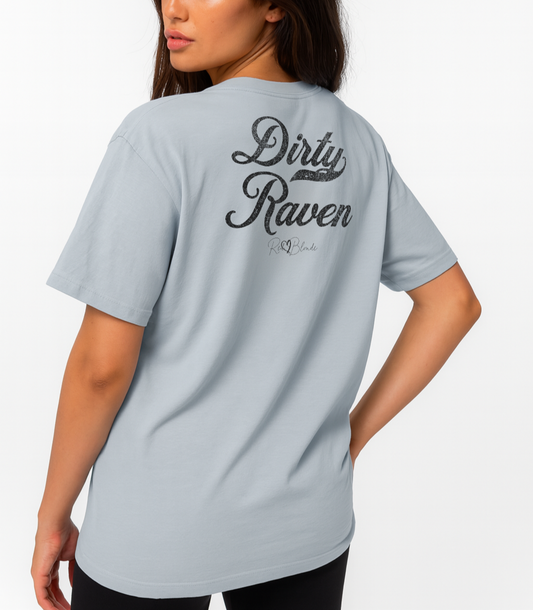 Raven haired woman wears pale blue vintage wash oversized unisex T-shirt with bold black Milkstore 01 vintage-style script reading “Dirty Raven” and the RedBlonde signature logo beneath, plus a small RedBlonde logo detail on the right sleeve.