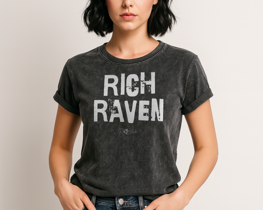 raven haired model wears a black acid-wash unisex denim T-shirt with a distressed vintage look. The shirt features the bold white text “RICH RAVEN” in a weathered Ed Gein font, and features the RedBlonde signature logo. The shirt has short sleeves, a crew neckline, and visible seam detailing for a rugged, casual style. Branded Redblonde on inside neck and right sleeve