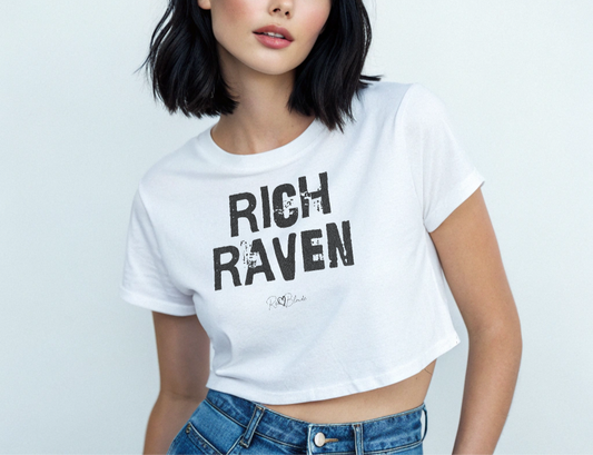raven haired model wears a white crop top with a relaxed fit, short sleeves, and a wide crew neckline. The front features bold black distressed lettering reading “RICH RAVEN” with the RedBlonde signature logo underneath in black