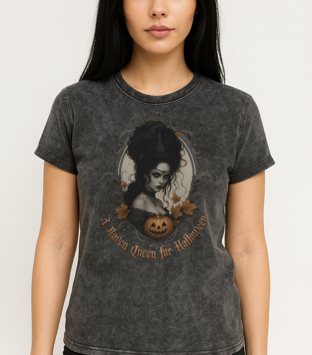 woman with black hairs wears a Black acid-wash unisex T-shirt featuring a gothic-style illustration of a dark-haired woman with an elaborate beehive hairstyle, framed by ornate scrollwork, autumn leaves, and a carved pumpkin. The design is captioned with the text ‘A Raven Queen for Halloween’ in vintage Dark Angle lettering. Branded RedBlonde in red on inside neck and right sleeve