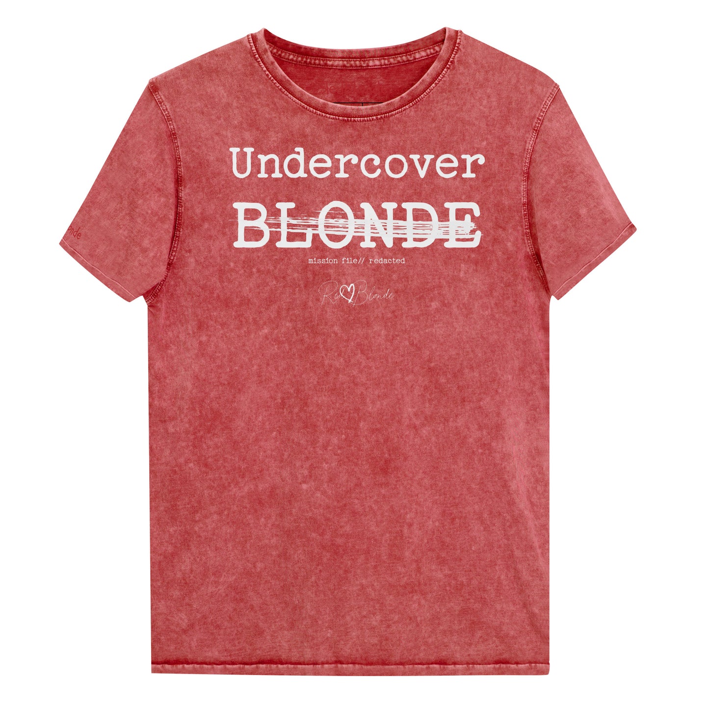 A red unisex denim-style t-shirt with a vintage washed finish. The front features bold white text reading “Undercover BLONDE” with “BLONDE” partially struck through by a horizontal white bar. Beneath it, smaller text says “mission file // redacted” along with a subtle RedBlonde logo. The shirt has a classic crew neck and short sleeves with visible stitching for a rugged look. Branded RedBlonde London in red on inside neck and right sleeve