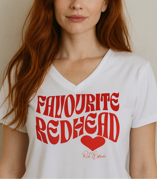 redhead wears a White women's loose fit v-neck graphic t-shirt with the slogan 'Favourite Redhead' in Solino Display red retro 70s wavy font. Signed with a heart and RedBlonde. Branded RedBlonde in red on inside neck and right sleeve