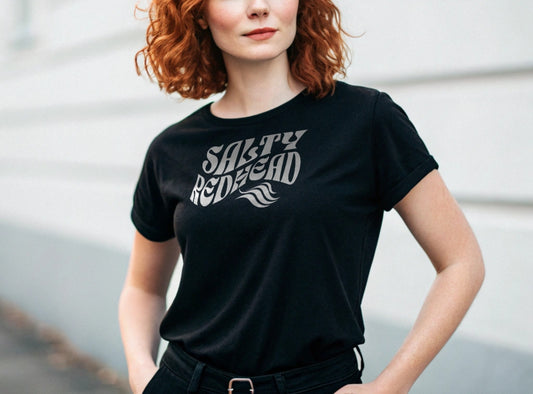 redhead wears a black classic unisex graphic t-shirt with the slogan 'Salty Redhead' in Solino Display white retro 70s wavy font. Branded RedBlonde in red on inside neck and right sleeve