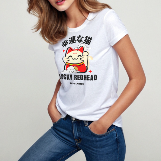 redhead wears a white unisex T-shirt featuring a colorful maneki-neko (lucky cat) graphic wearing a red outfit with gold accents. Above the cat, Japanese kanji characters are printed in bold white. Below the illustration, the text reads “LUCKY REDHEAD” in distressed white font, with “REDBLONDE” in smaller uppercase letters underneath. The shirt has short sleeves, a classic crew neckline, and a casual, modern fit.