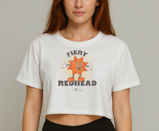 redhead wears White women’s crop top with a bold retro graphic of a smiling orange cartoon sun character, surrounded by radiant sunbeams. The text reads ‘Fiery Redhead’ in distressed black Belzebuth lettering with small details including ‘ESTD 1994’ and the RedBlonde signature logo beneath."