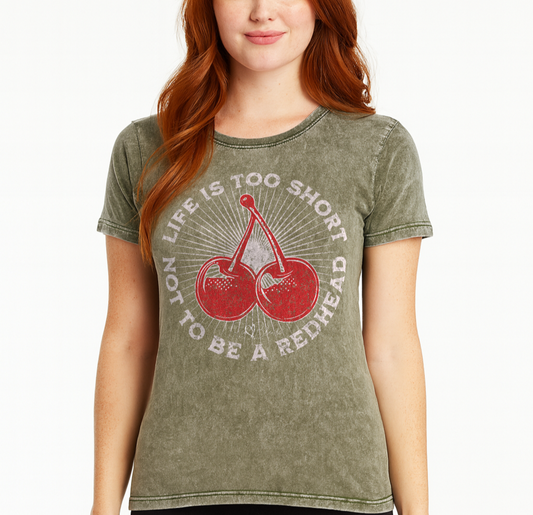 Redhead wears a Dark army green acid-wash unisex T-shirt featuring a bold red cherry graphic in the center. Surrounding text reads “Life is too short not to be a redhead” in distressed white adventure font lettering. The inside neck and sleeve also feature the RedBlonde logo in red.