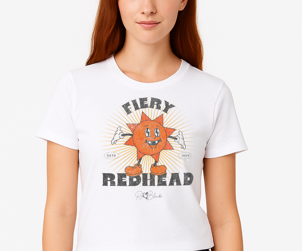redhead wears a White unisex crew neck t-shirt with a bold retro graphic of a smiling orange cartoon sun character, surrounded by radiant sunbeams. The text reads ‘Fiery Redhead’ in distressed black Belzebuth lettering with small details including ‘ESTD 1994’ and the RedBlonde signature logo beneath."