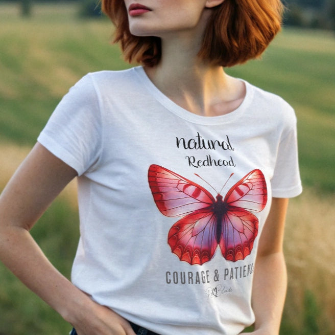 redhead woman wears white unisex relaxed fit t-shirt with the words natural redhead in arionna font above a graphic of a red butterfly with 'COURAGE & PATIENCE' below in adventurer 06 font signed off by RedBlonde.