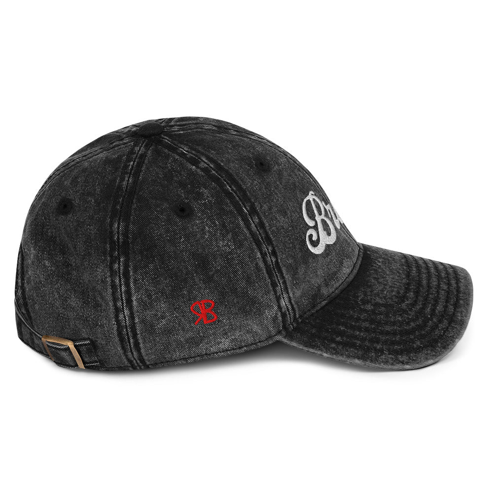 Flatlay side view of black denim vintage wash effect baseball cap with the word 'Brunette' embroidered on the front in white adventure retro font with the initials RB in red which stand for the brand RedBlonde