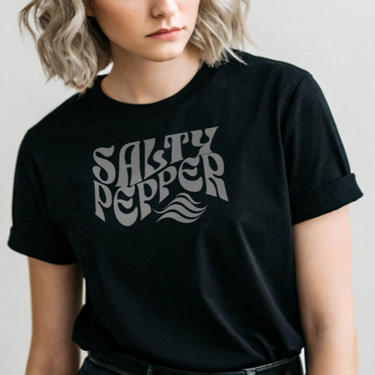 silver haired woman wears a black classic unisex graphic t-shirt with the slogan 'Salty Pepper' in Solino Display white retro 70s wavy font. Branded RedBlonde in red on inside neck and right sleeve
