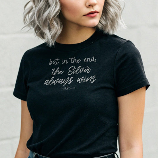 silver haired youthful woman wears a black unisex t-shirt featuring the handwritten-style slogan “but in the end, the Silver always wins” in Hillbear font in dark gray text across the chest, signed off RedBlonde. Branded “RedBlonde London on inside the collar and on the right sleeve