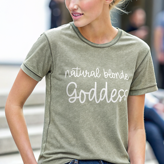 Blonde model, sits on steps, wearing jeans and a unisex Army Green Vintage denim wash graphic tee with “Natural Blonde Goddess” in handwritten text in white