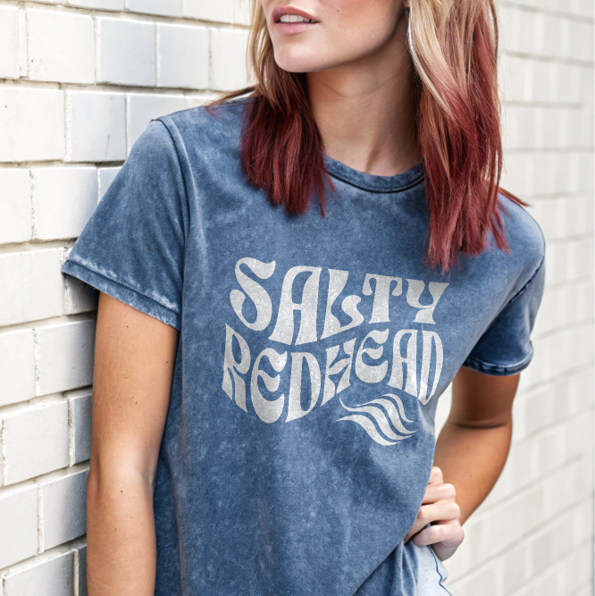 redhead model by wall wears jean shorts and a unisex relaxed fit denim-wash t-shirt with the words 'Salty redhead' in retro 70s vibe text in white.