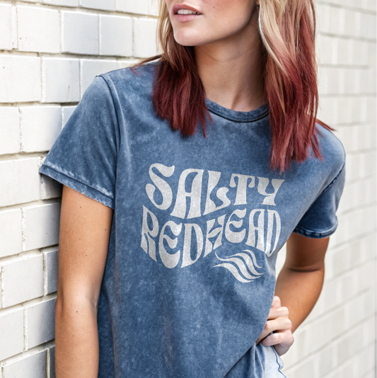 redhead model by wall wears jean shorts and a unisex relaxed fit denim-wash t-shirt with the words 'Salty redhead' in retro 70s vibe text in white.