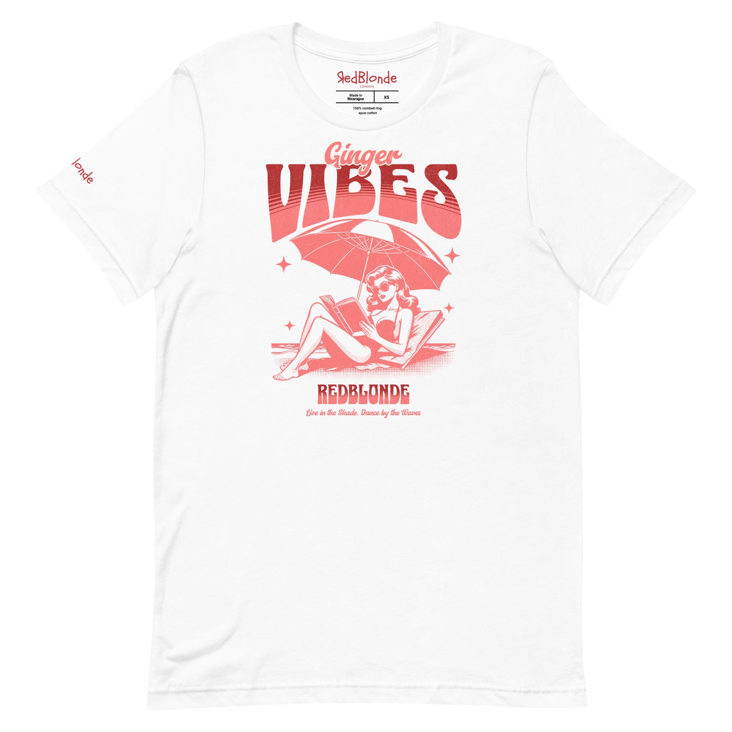 white unisex t-shirt featuring a retro-style red illustration of a redhead woman lounging under a beach umbrella reading a book, with bold text reading "Ginger Vibes" above and "RedBlonde" below, alongside the tagline "Live in the shade. Dance by the waves." Branded RedBlonde on inside neck and right sleeve