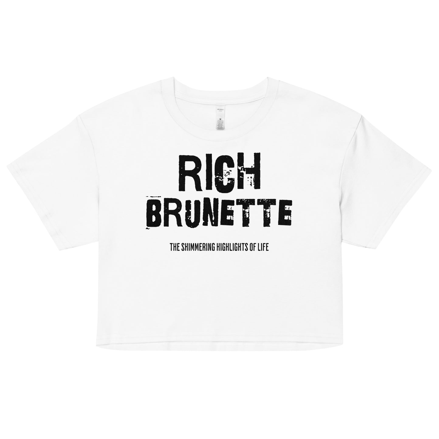 A white women’s crop top with a relaxed fit, short sleeves, and a wide crew neckline. The front features bold black distressed lettering reading “RICH BRUNETTE” with the tagline “THE SHIMMERING HIGHLIGHTS OF LIFE” in smaller text below.